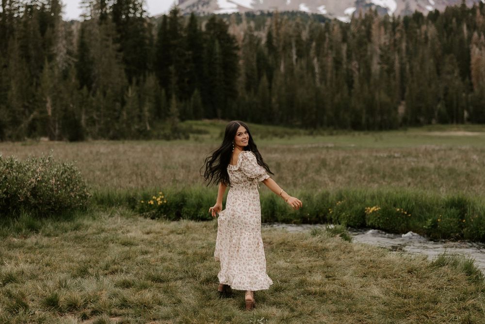A person in a floral maxi dress twirls in a mountain meadow with evergreen forest and snow-capped peaks in the background.