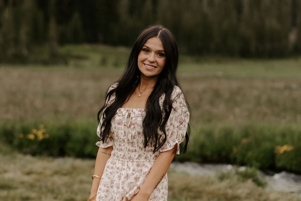 A woman in a floral dress poses outdoors in a natural setting with grass and trees in the background.