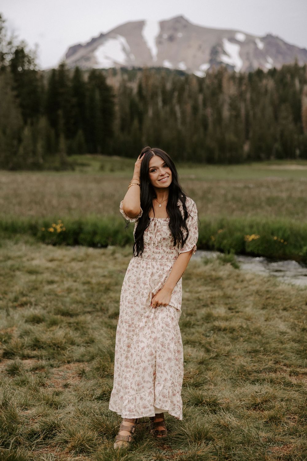A person in a floral maxi dress poses in a mountain meadow with snow-capped peaks and evergreen forest in the background.