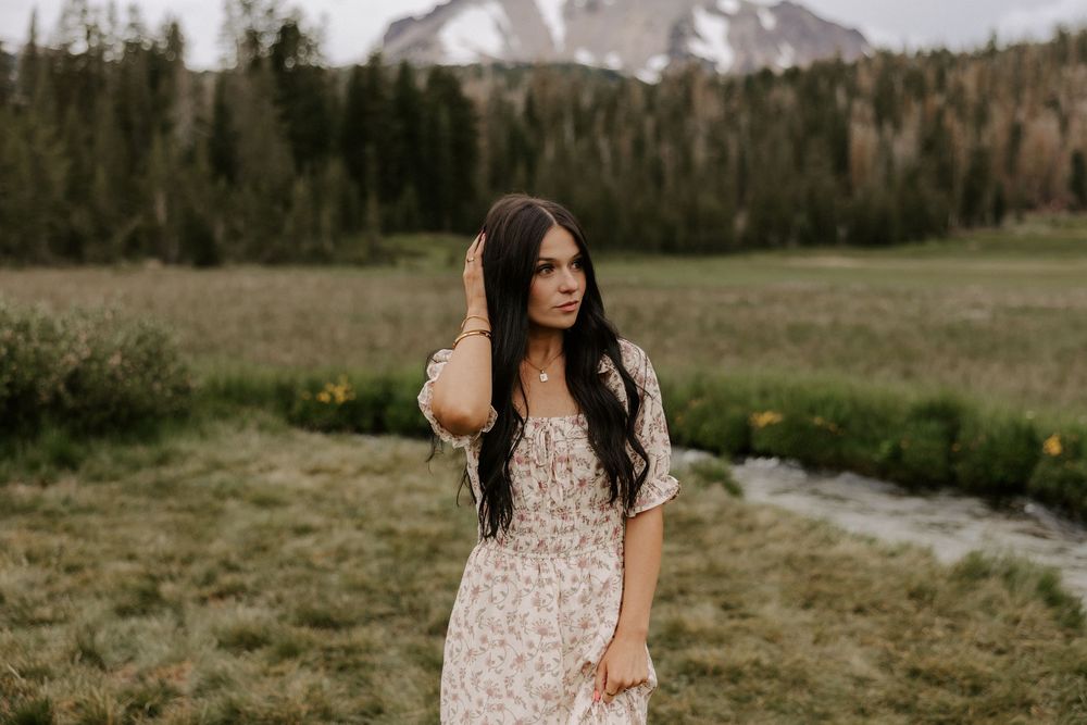 A person in a floral dress stands in a scenic mountain meadow with evergreen trees and snow-capped peaks in the distance.
