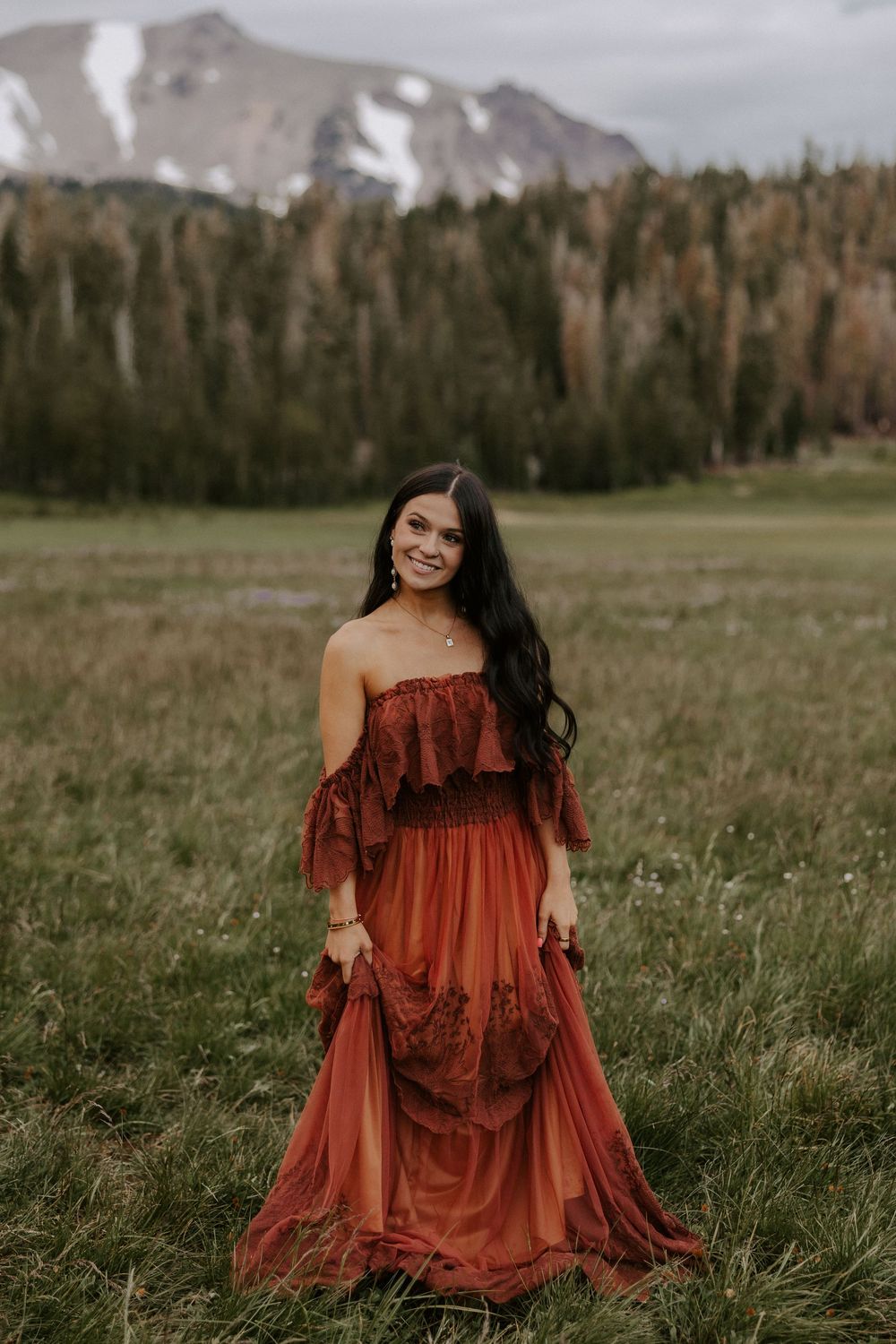 A woman in a rust-colored dress sits gracefully in a meadow with mountains and forest in the background.