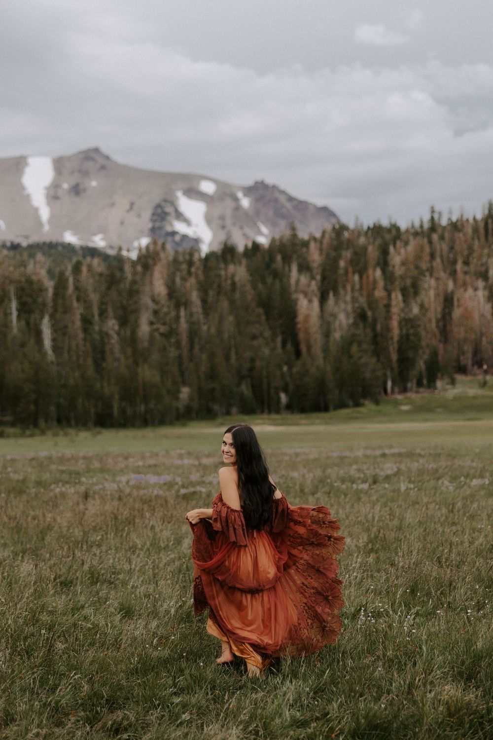 A flowing rust-colored dress creates movement against a mountainous landscape with evergreen trees.