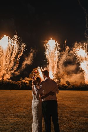 A couple watches a spectacular fireworks display in the distance across a grassy field at dusk.