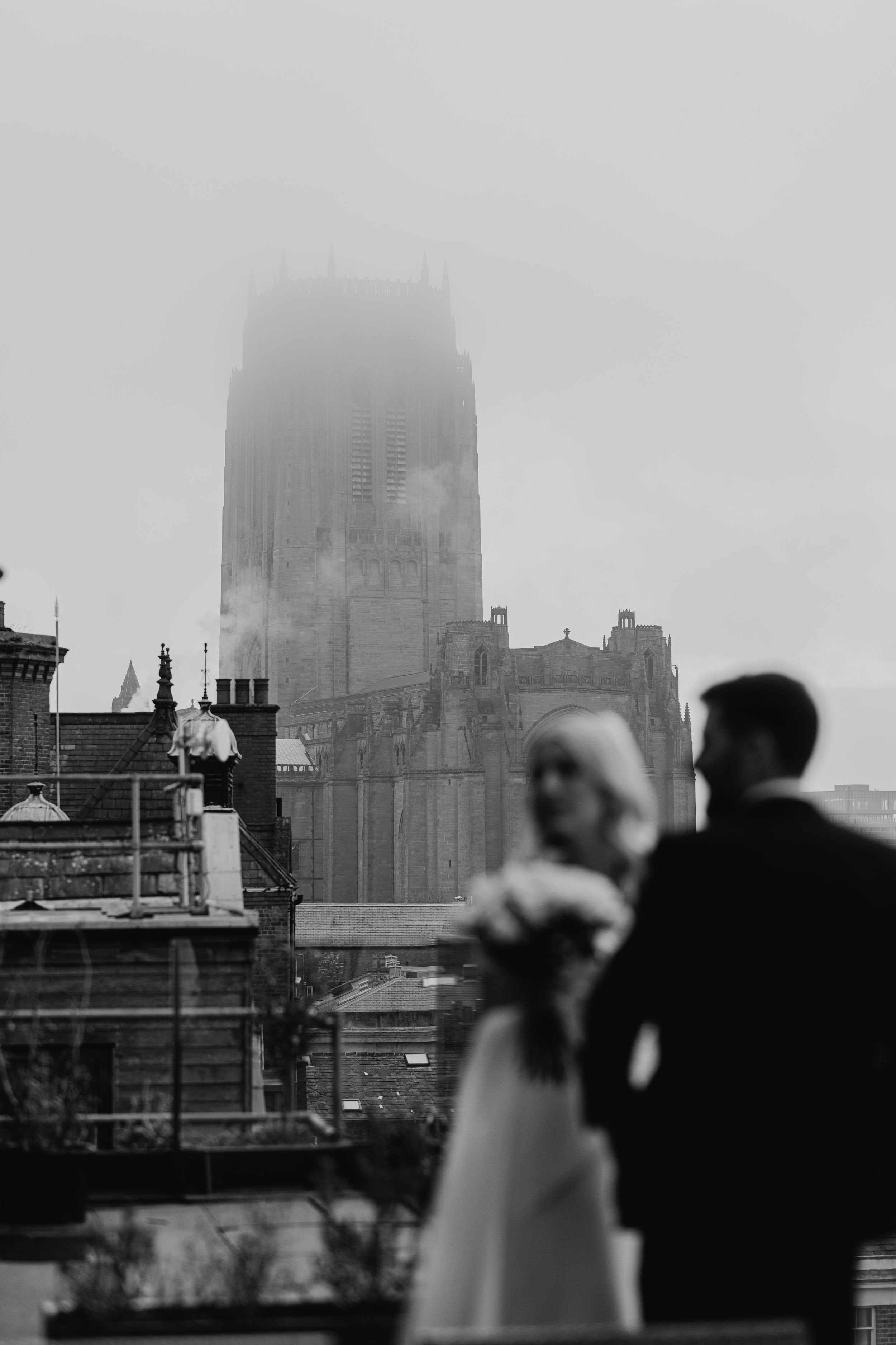 A moody black and white cityscape with Liverpool Cathedral appearing misty in the background as silhouettes stand in front.