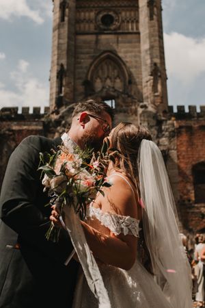 A romantic wedding couple shares a passionate kiss outside a historic stone church on their special day.
