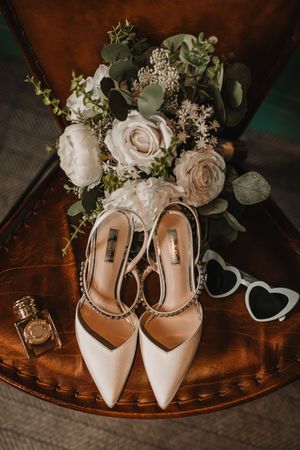 Elegant white bridal shoes with floral bouquet arrangements displayed on a vintage wooden surface in warm lighting.