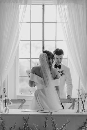 Black and white wedding photo of a couple sharing an intimate moment while cutting their cake by a sunlit window.