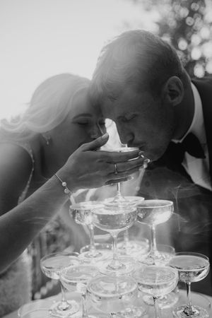A black and white artistic photo shows a champagne tower being created with multiple coupe glasses in soft lighting.