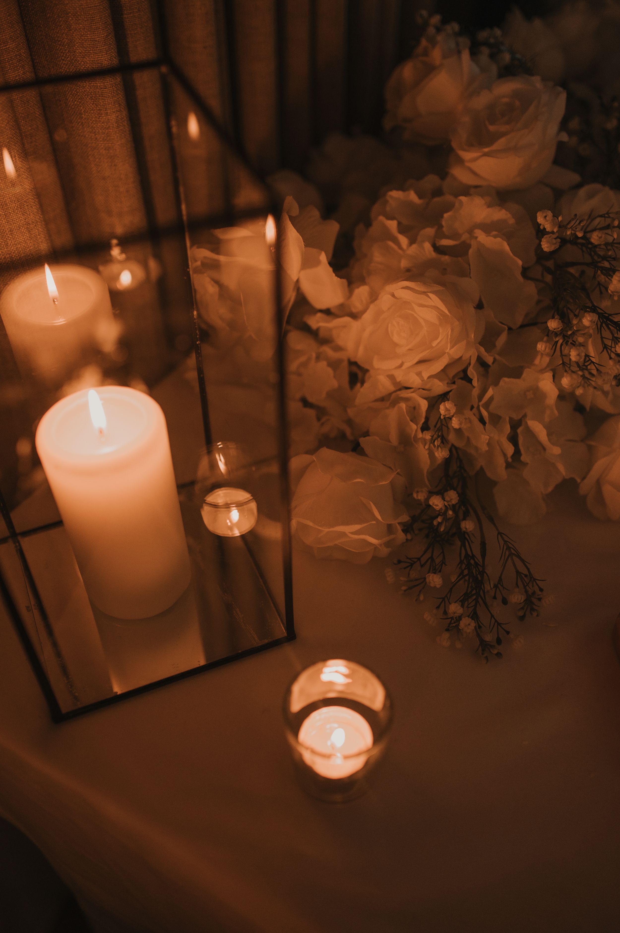 Lit candles and white roses create a romantic ambiance in warm sepia tones with mirrors reflecting the soft glow.