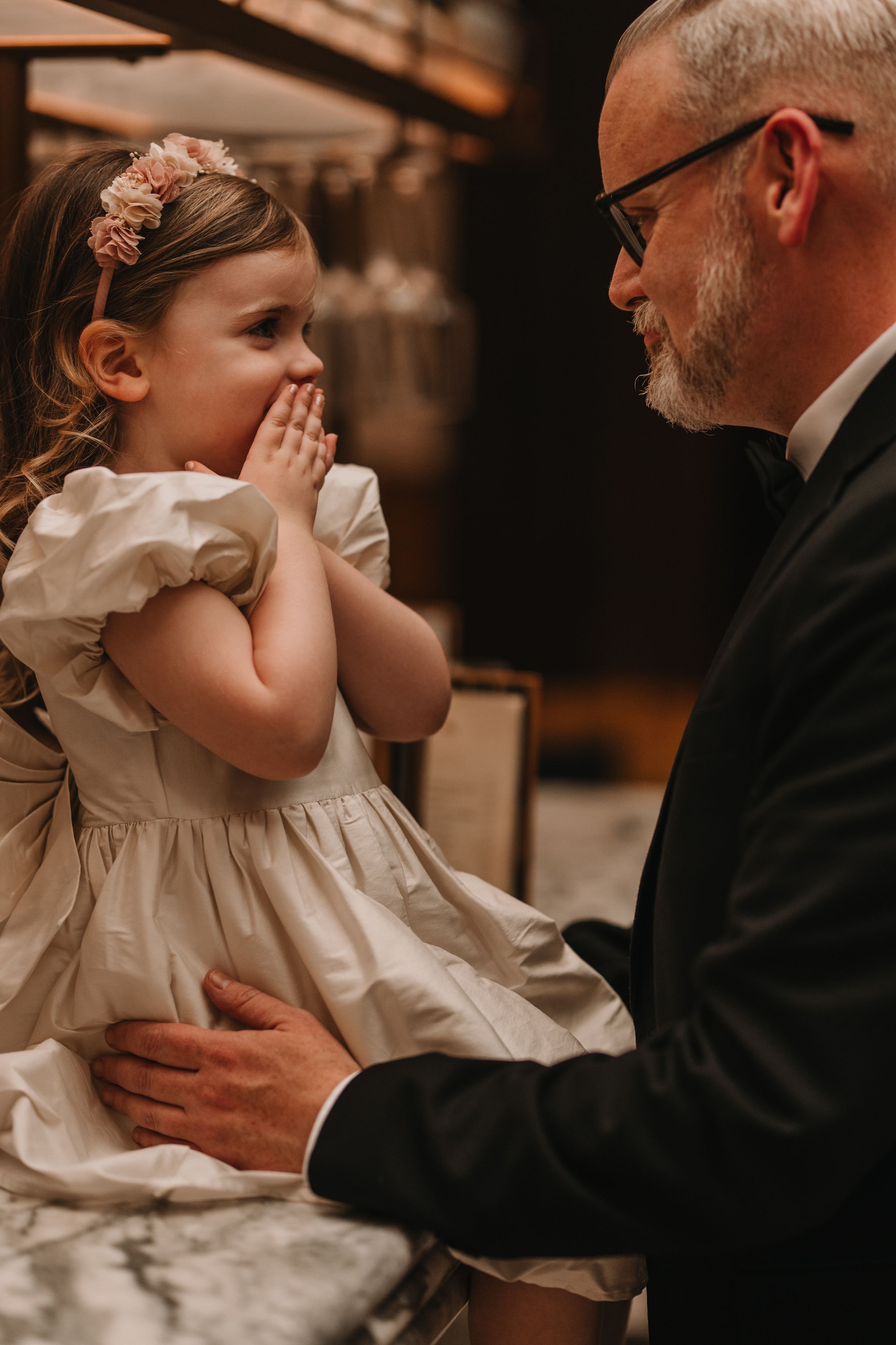 A heartwarming candid moment between a young girl in a cream dress and floral headband with someone in formal attire.