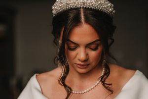 A bride wearing a delicate pearl tiara and necklace poses with closed eyes in a romantic bridal portrait.