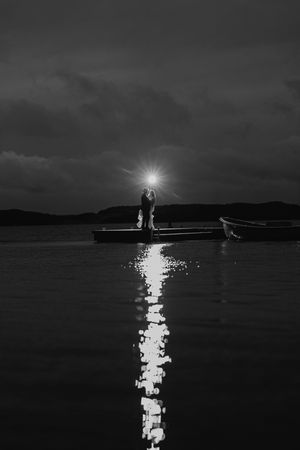 A silhouetted couple stands on a dock at night with moonlight creating a glowing reflection on the dark water.