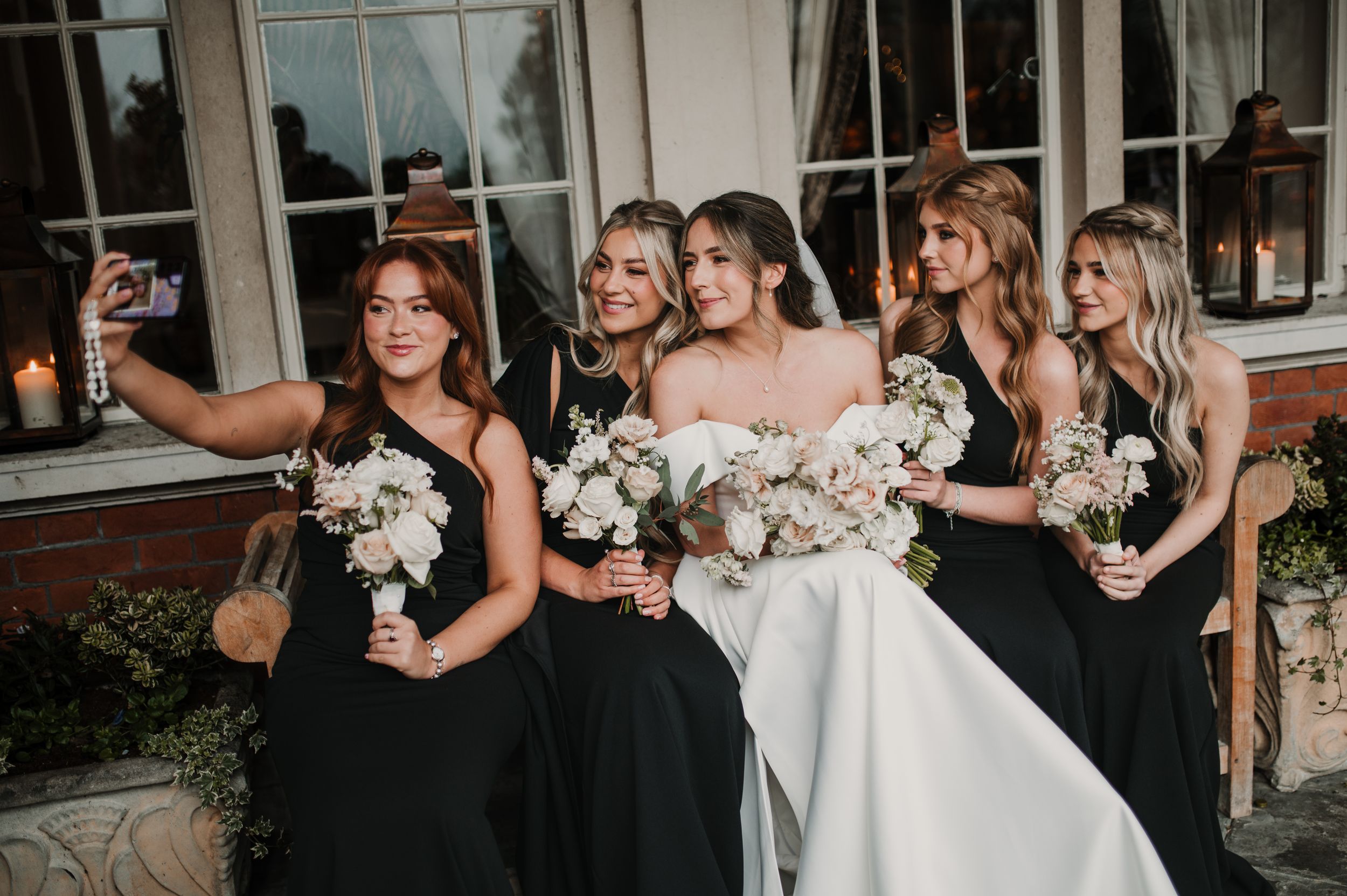 Wedding party in black dresses poses together with bride in white gown holding floral bouquets.