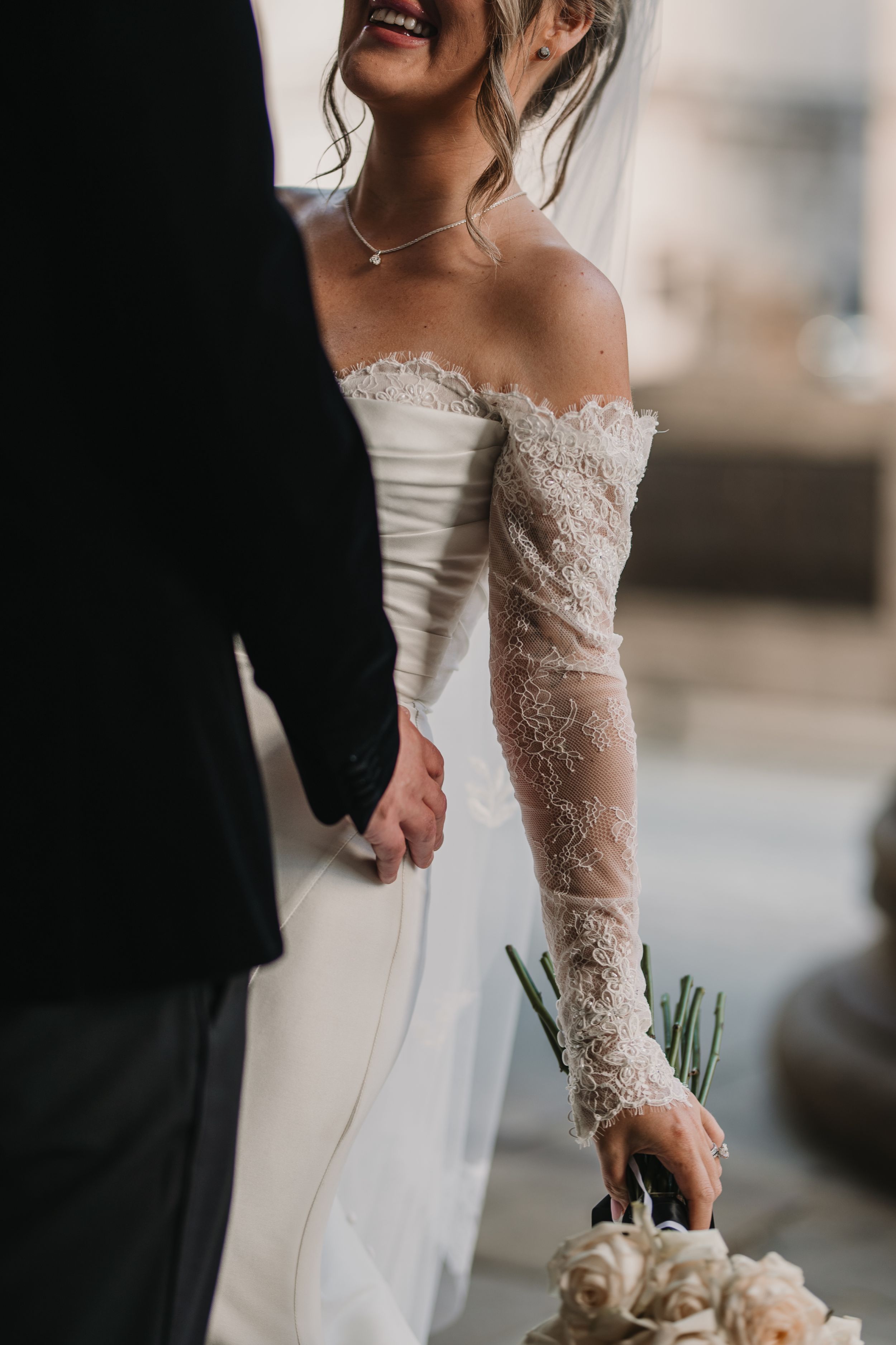 Close-up detail of bride's lace sleeve and white bouquet as she holds hands with groom in formal wedding attire.