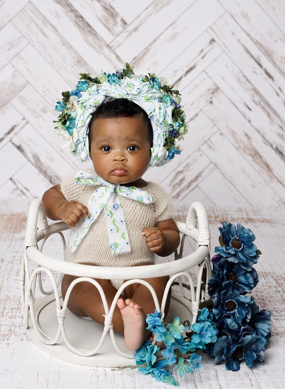 baby girl posed in front of wooden wall seated in a basket surrounded by teal flowers wearing a teal floral bonnet.