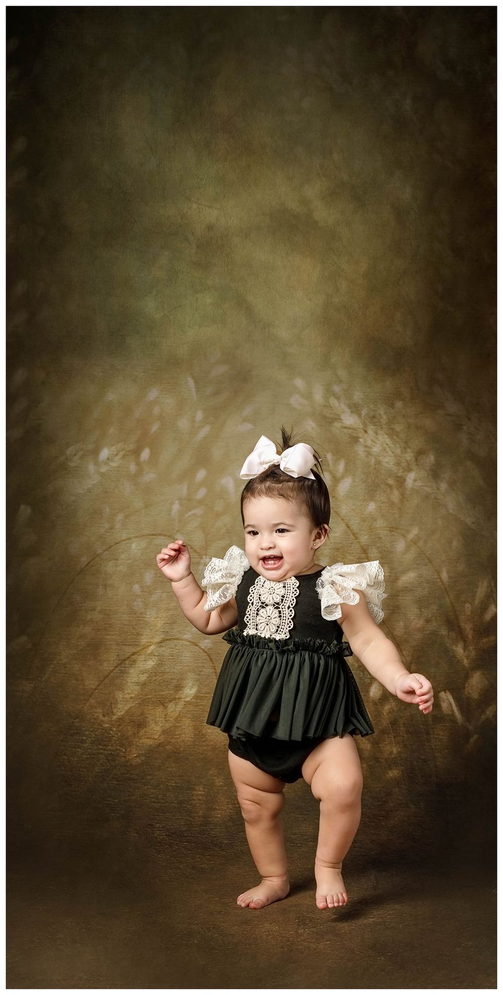 baby girl in a olive green and cream dress standing, taking first steps in front of a natural toned background