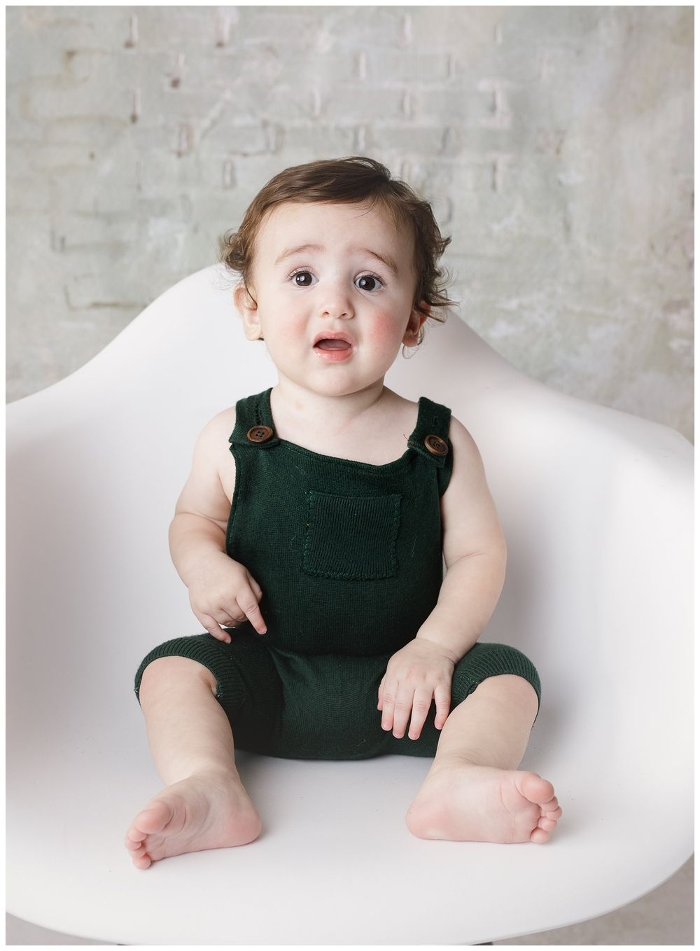 adorable 9 month old boy posed in front of whitewashed brick wall wearing forest green overalls.