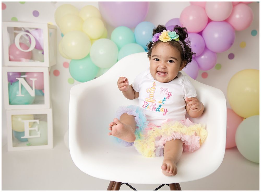 little girl in pastel rainbow in front of matching balloon arch and blocks that spell out ONE.
