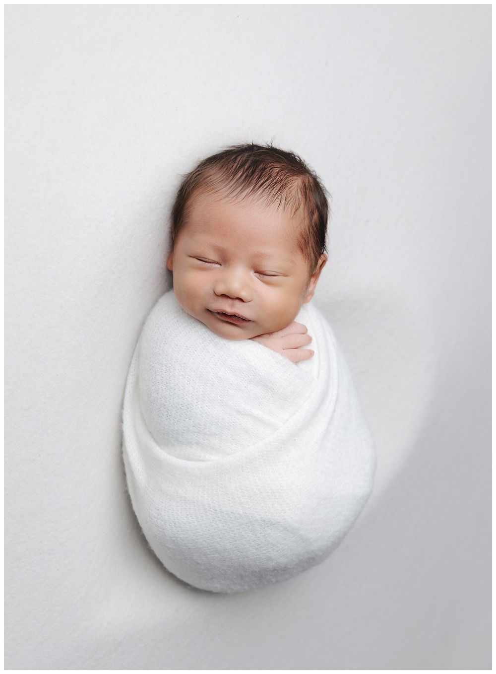 newborn boy, swaddled posed in a heart shape background. swaddled in white on a white backdrop.