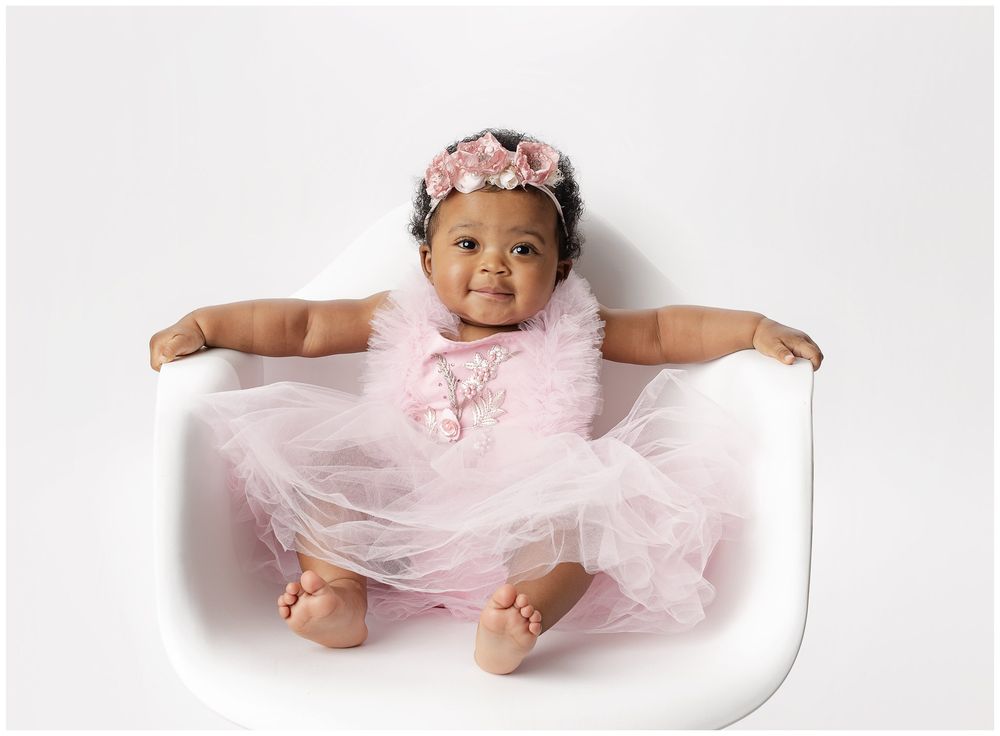 baby girl on her first birthday in a pink tulle dress, sitting in a white plastic modern chair on a white background.