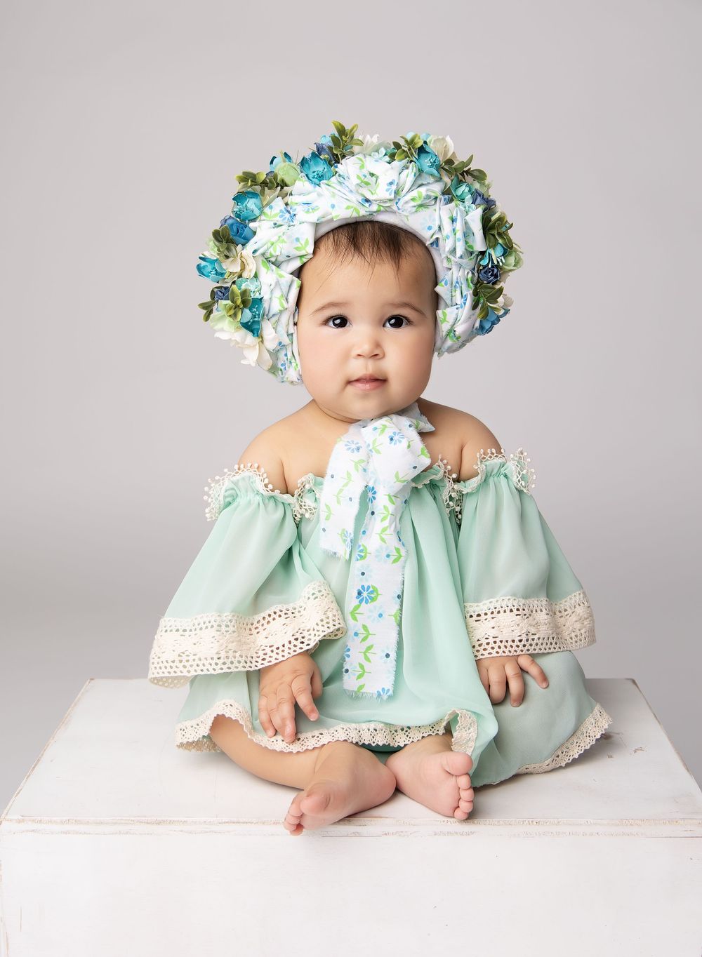sitting baby in a mint green dress with flowy sleeves and large, oversized flower bonnet.