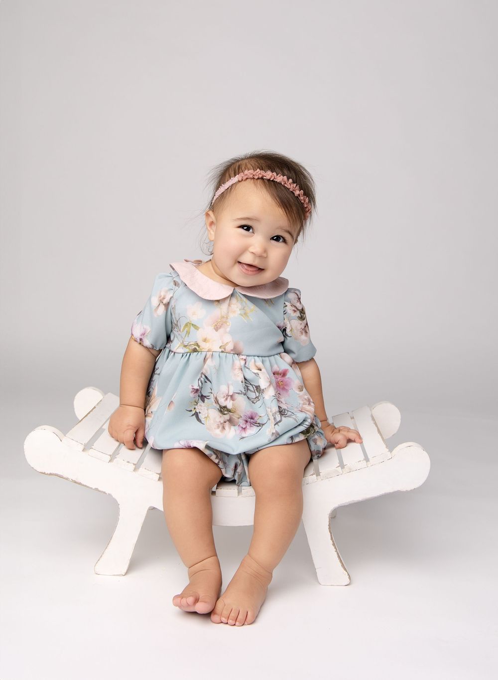 Adorable baby girl posed sitting on a curved bench wearing a light blue bubble romper with light pink floral design and peter pan collar.