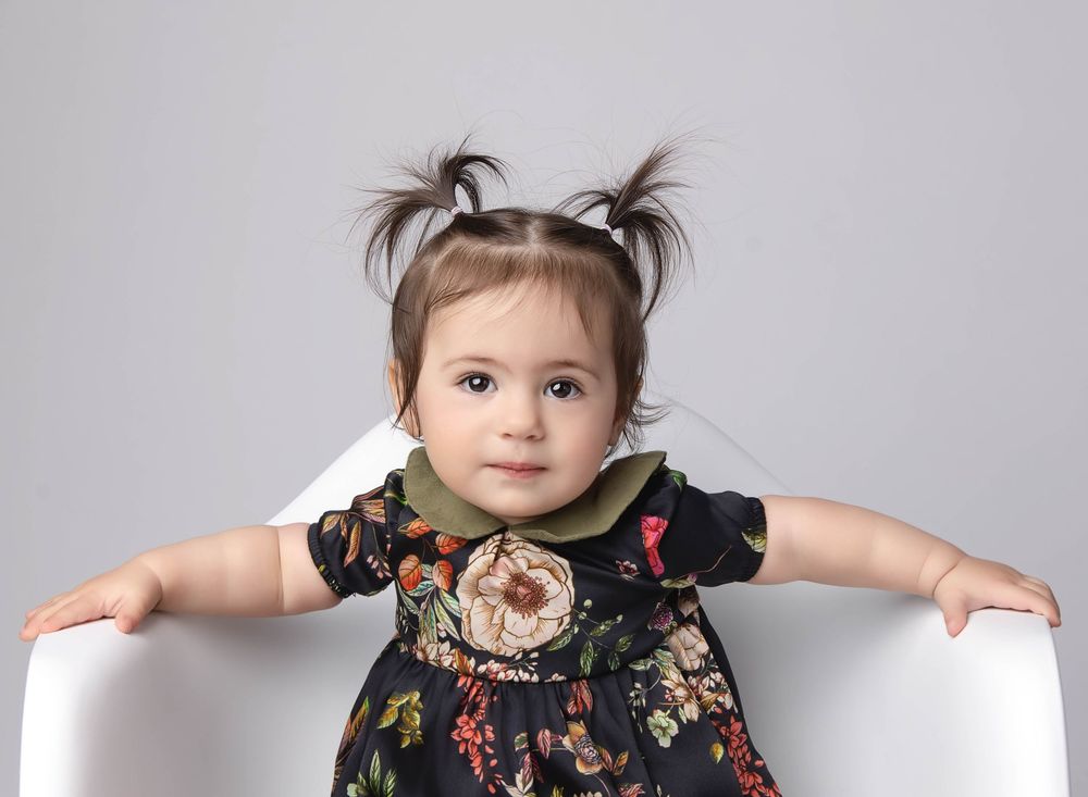 one year old girl in a black floral bubble romper with pigtails posed in an oversized white plastic mid century mod chair