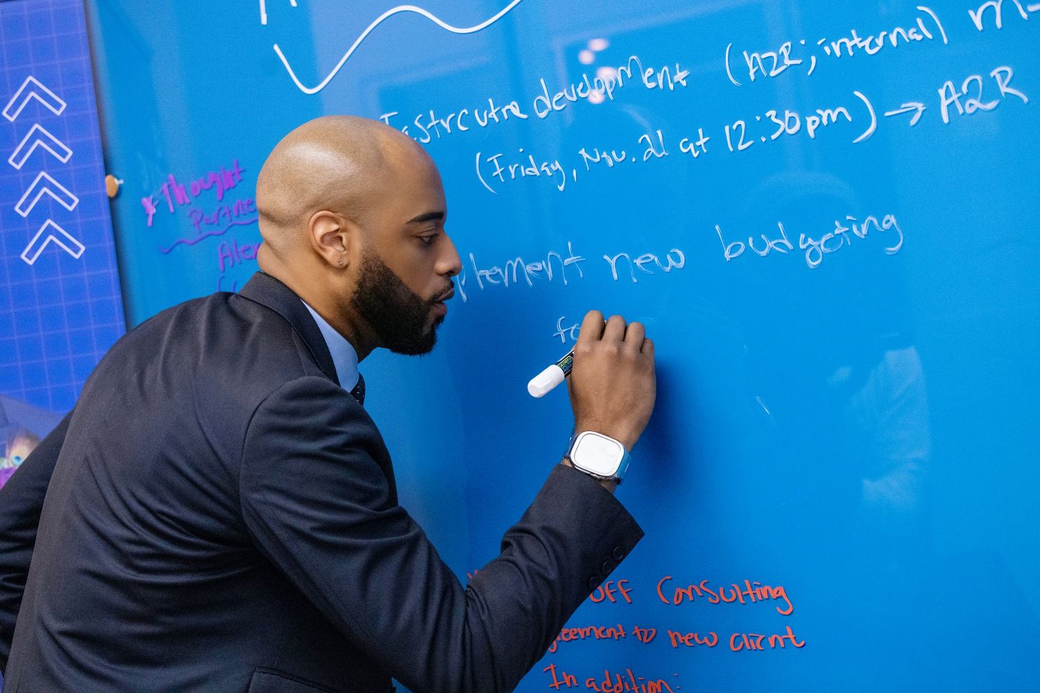 Someone writes mathematical equations and formulas on a bright blue wall-mounted whiteboard.