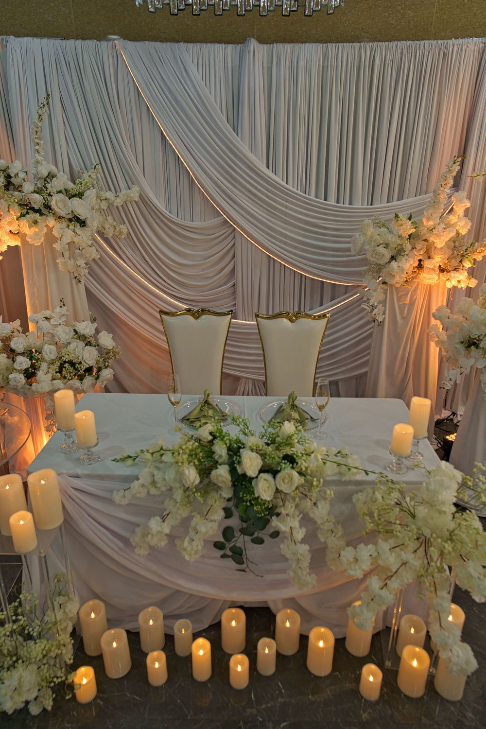 Elegant wedding reception table setup with white floral arrangements, draped fabric backdrop, and glowing pillar candles.