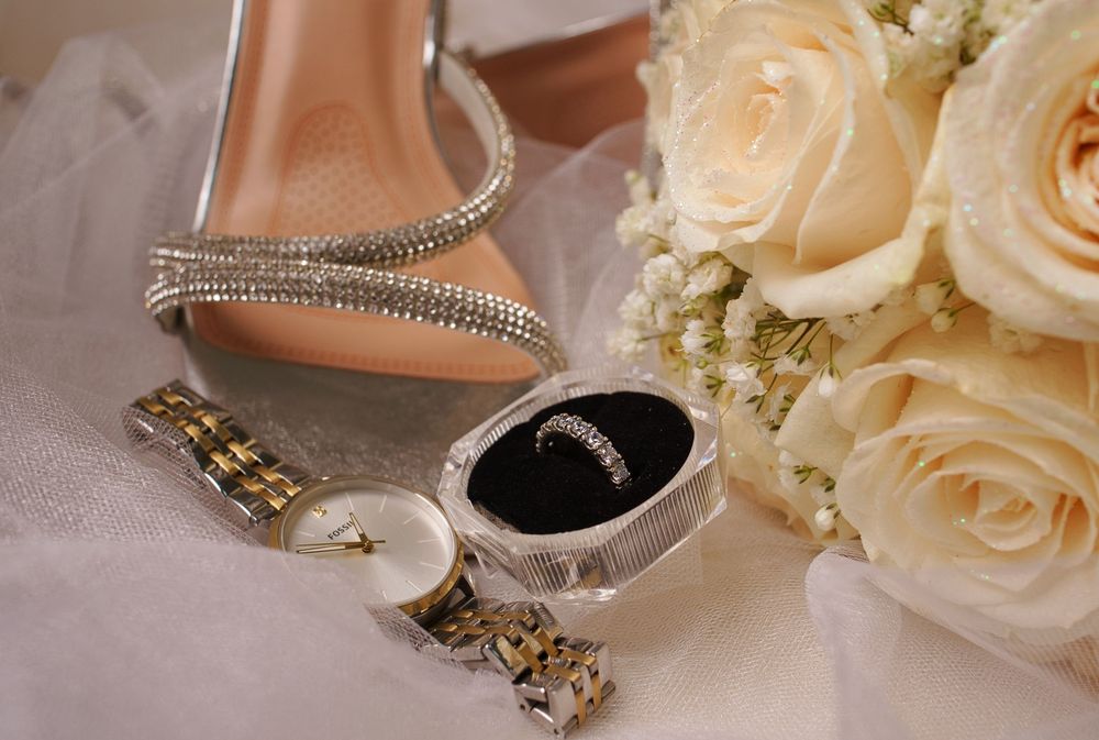 Wedding detail shot of delicate jewelry, rings, and cream roses arranged on a light colored surface.