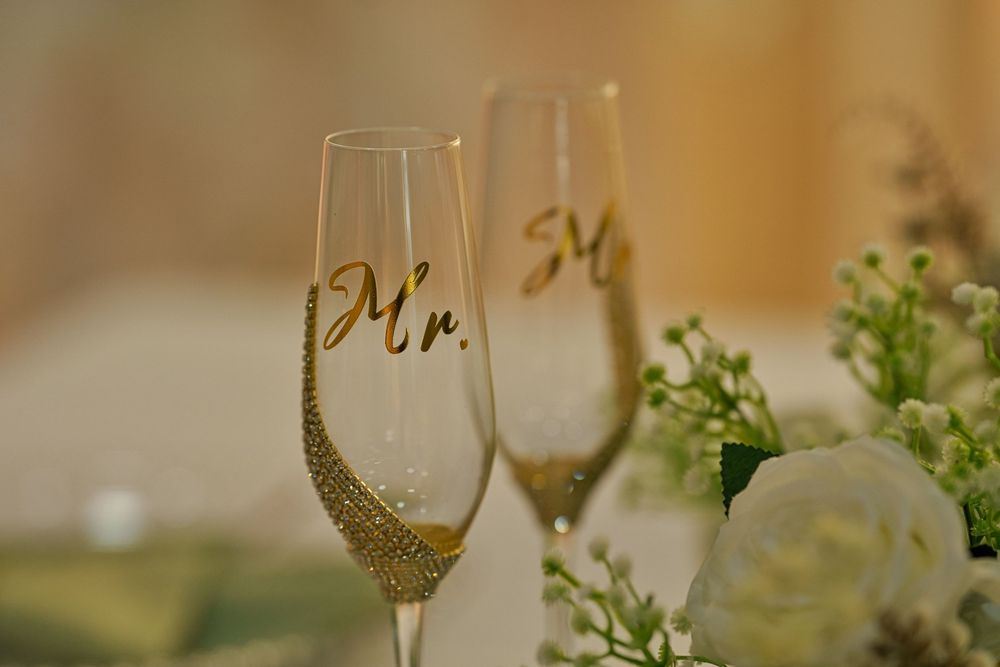 Two elegant champagne flutes decorated with gold Mr and Mrs text sit beside white roses and greenery in a wedding setting.