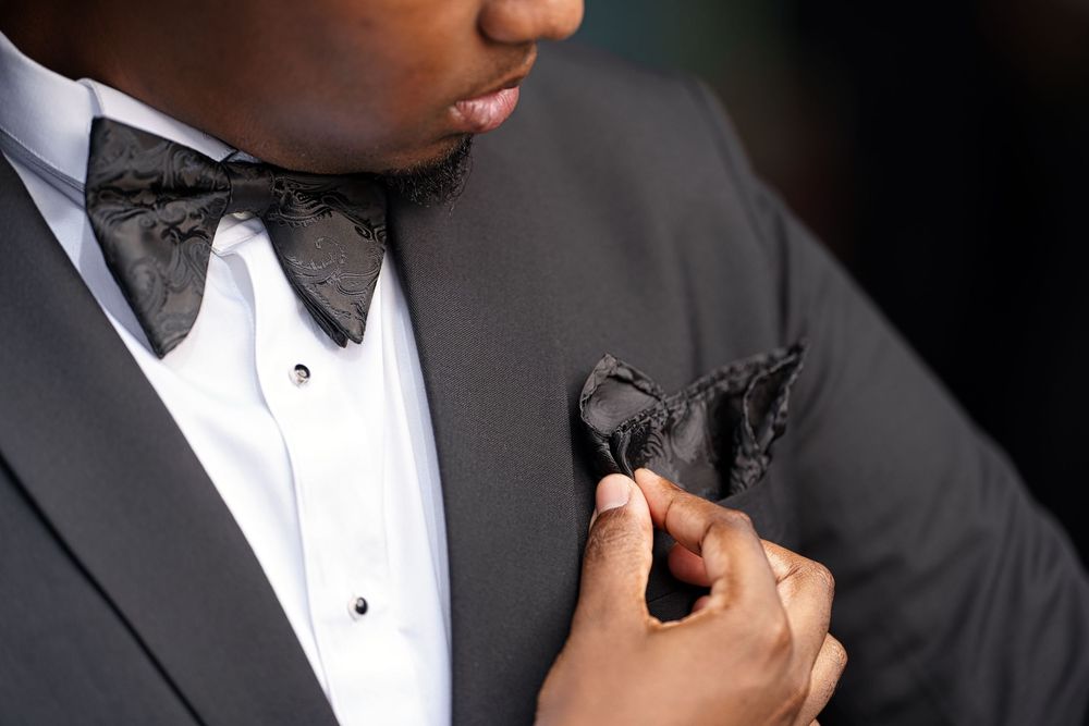 Someone adjusts a black bow tie while wearing a formal tuxedo with white dress shirt in a dark setting.