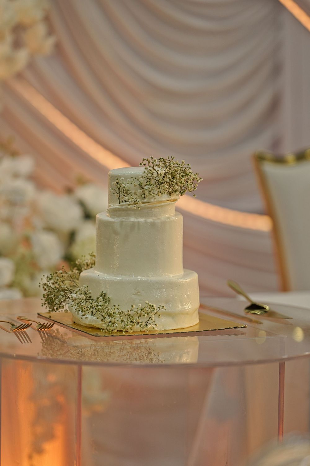 White wedding cake decorated with delicate baby's breath flowers sits on a clear acrylic stand with warm lighting.