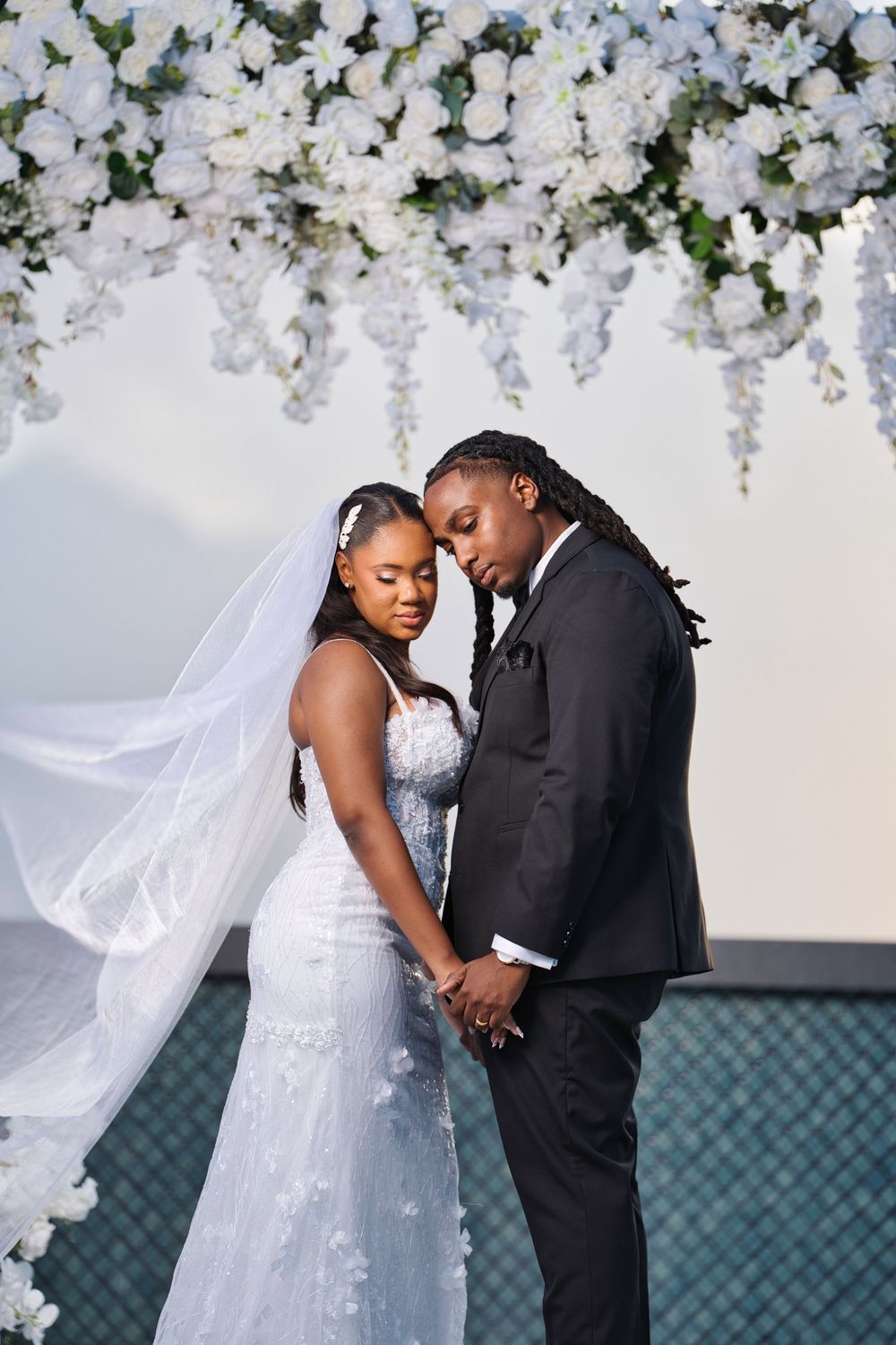 A romantic wedding couple shares intimate moments under white floral decorations against a teal backdrop.