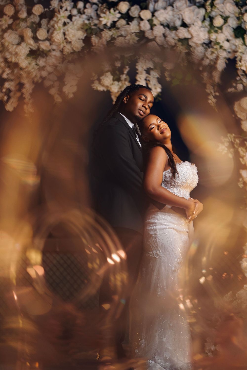 A romantic wedding couple embraces under soft floral blossoms with warm lighting creating a dreamy atmosphere.
