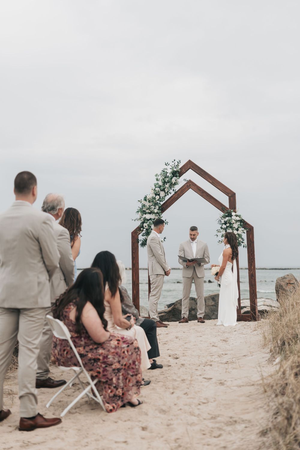 Intimate beach wedding ceremony with couple at wooden arch adorned with white flowers, guests watching by the ocean.