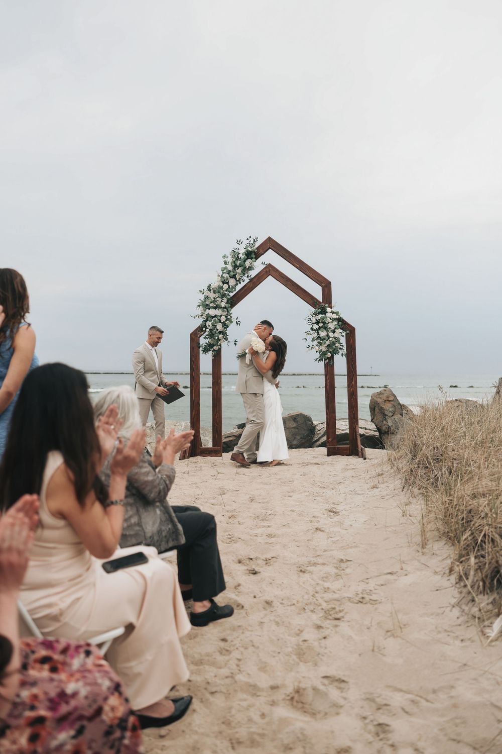 Couple shares first kiss under wooden arch with flowers on sandy beach wedding ceremony with guests applauding.
