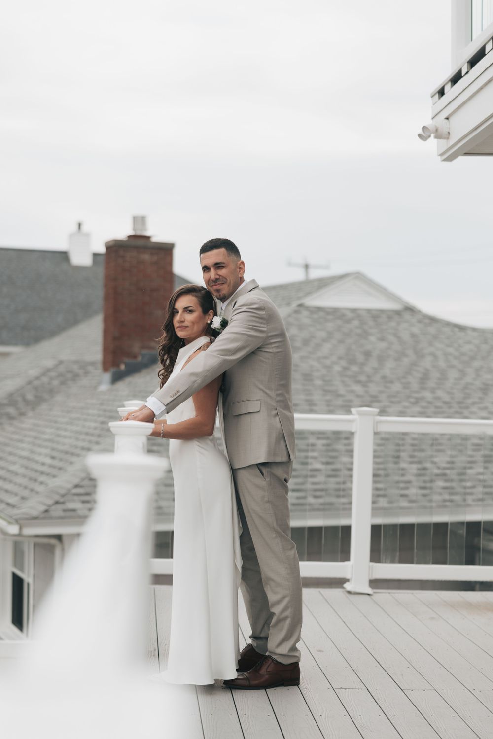 Bride and groom embrace on a rooftop deck, wearing white gown and tan suit, with gray shingled rooftops behind them.