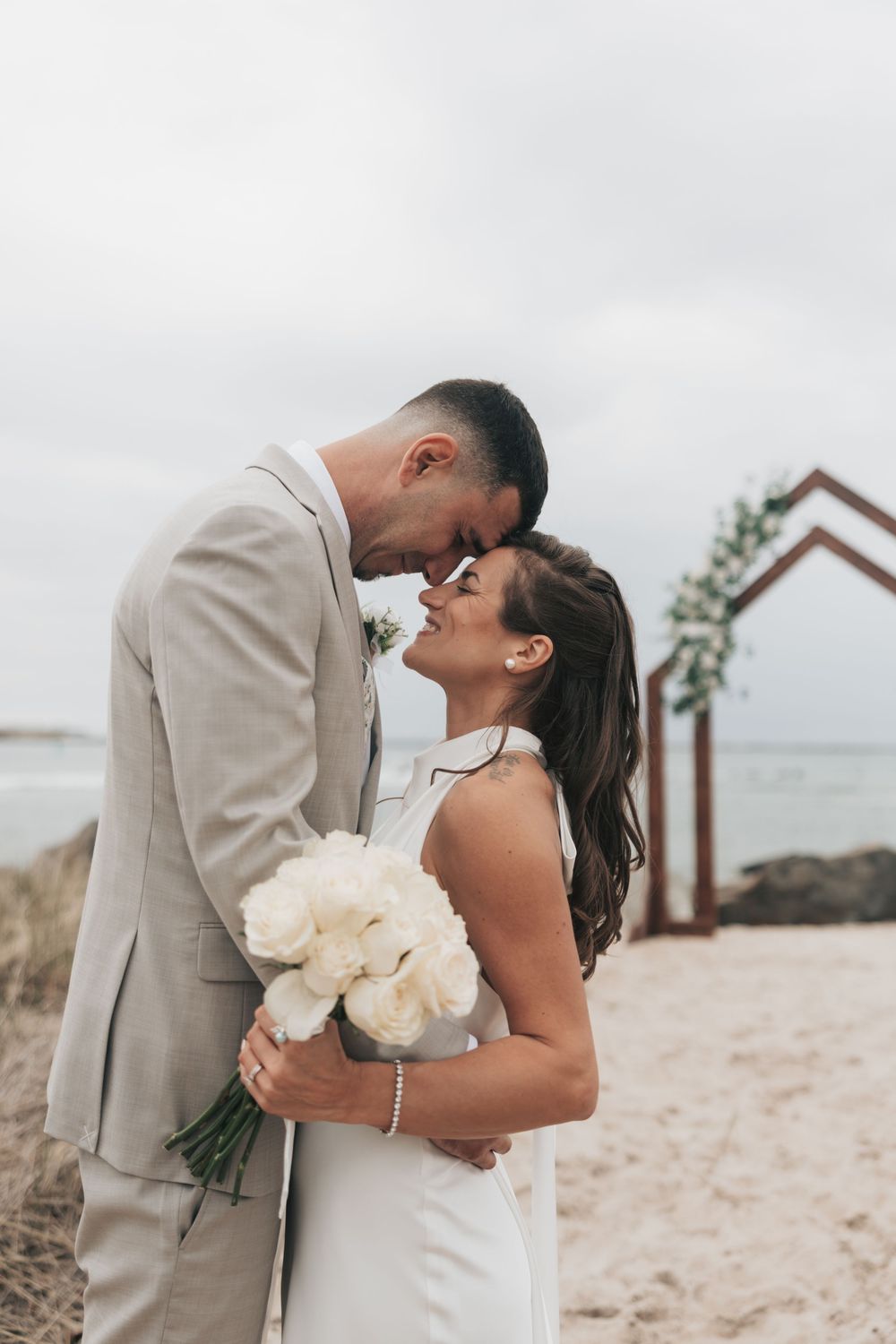 Bride and groom share a romantic forehead touch on a beach, she holds white peonies in a white gown and he wears a tan suit.