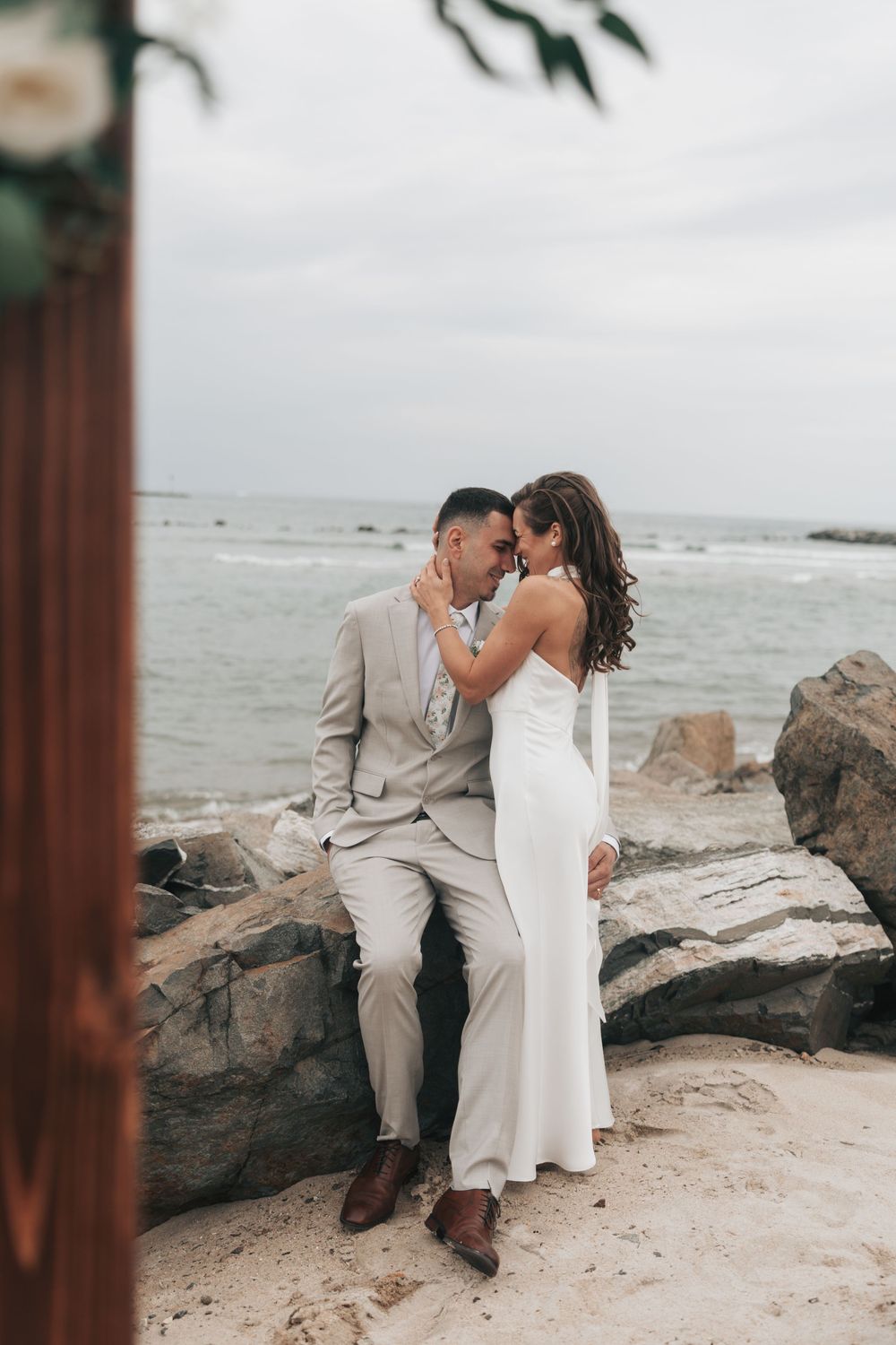 Bride and groom share an intimate moment on coastal rocks, she in a white gown and he in a tan suit with the ocean behind.
