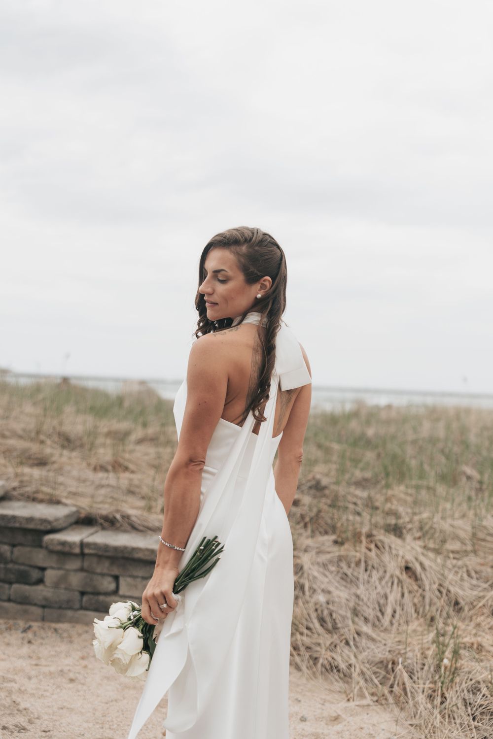Bride in white one-shoulder gown holding white bouquet on sandy beach, looking over shoulder on cloudy day.