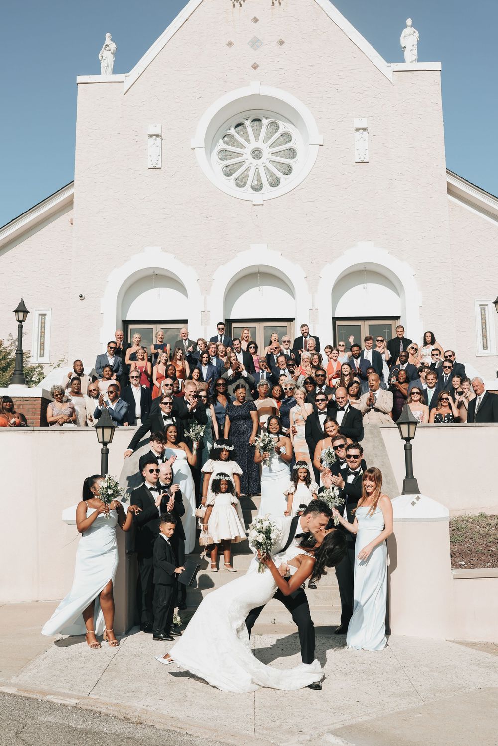 Bride and groom share a romantic dip kiss on church steps surrounded by wedding party and guests.