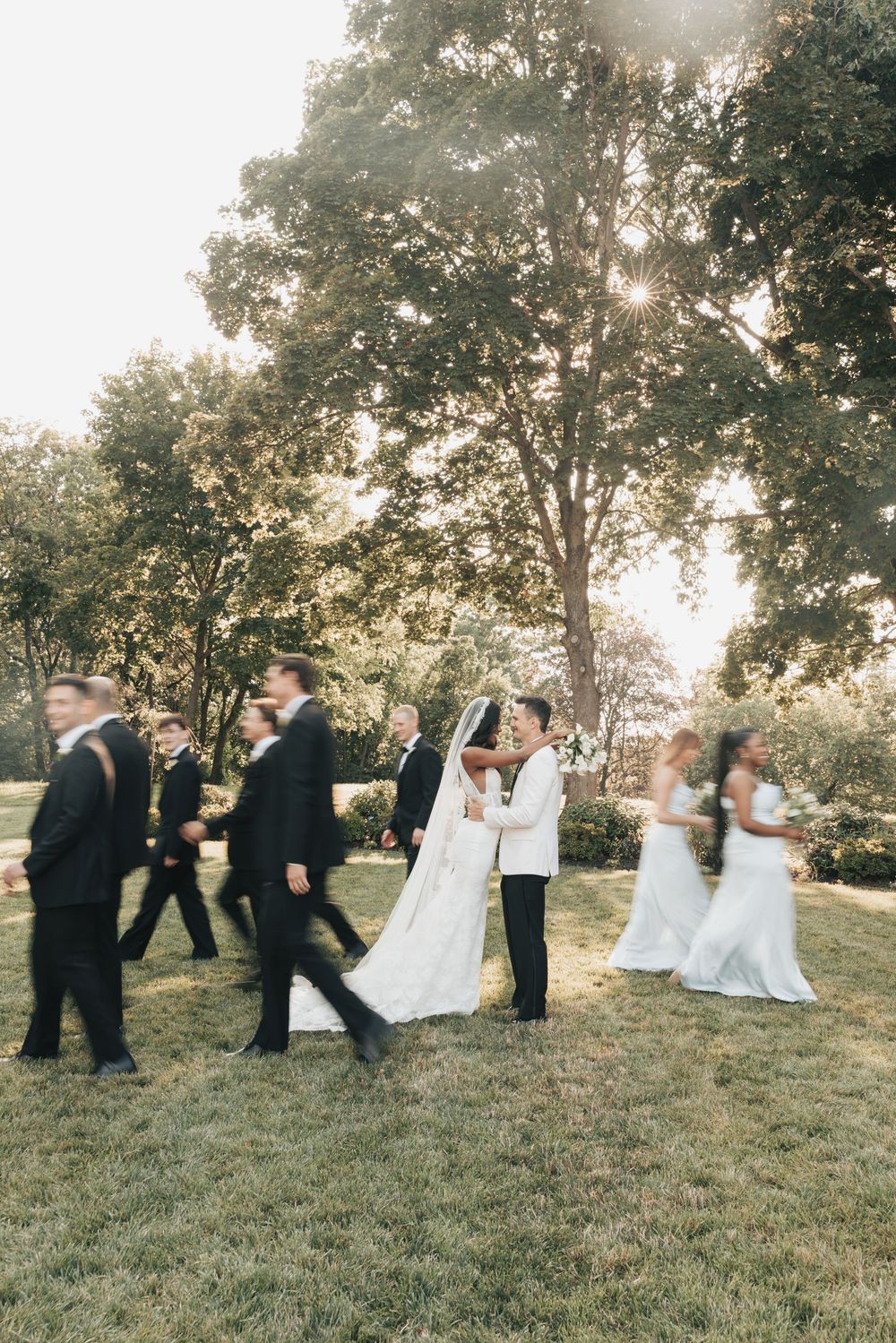 Bride and groom share a kiss surrounded by wedding party on lush green lawn with sunlit trees in background.
