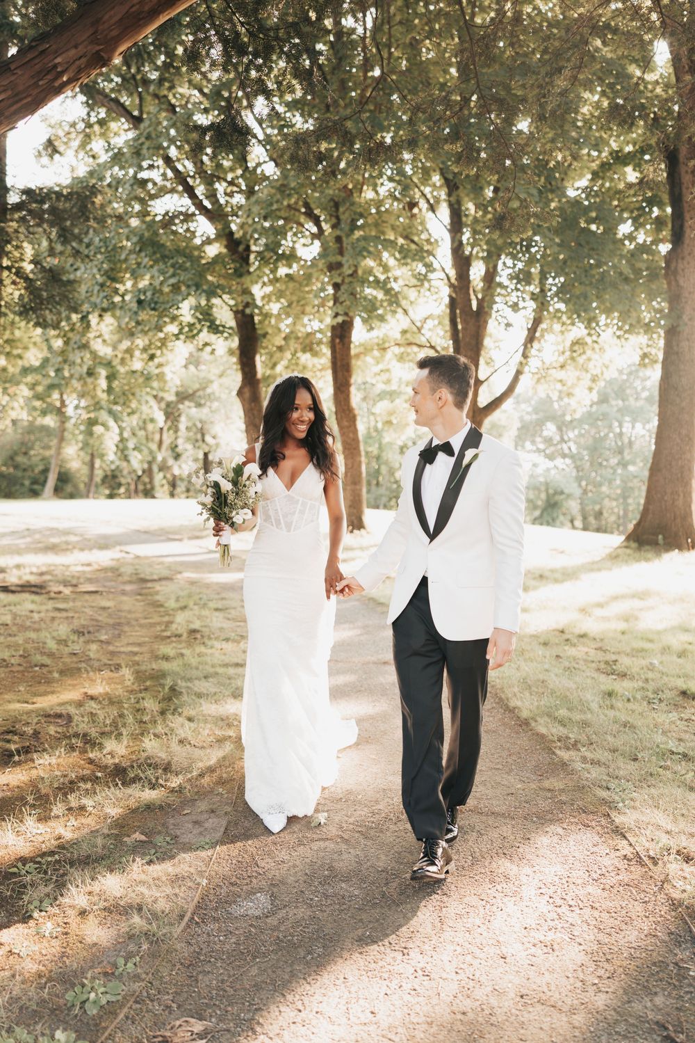 A bride and groom walk hand-in-hand along a sunlit path surrounded by lush green trees on their wedding day.