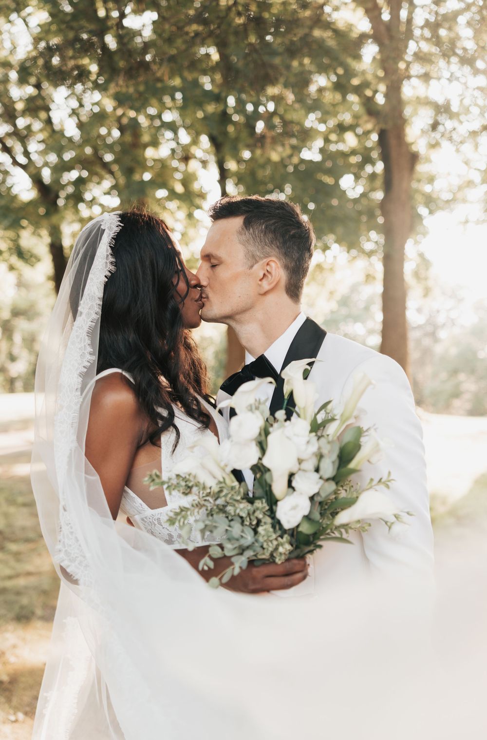 Bride and groom sharing a kiss outdoors, bride holding white floral bouquet with greenery, wearing wedding dress and veil.