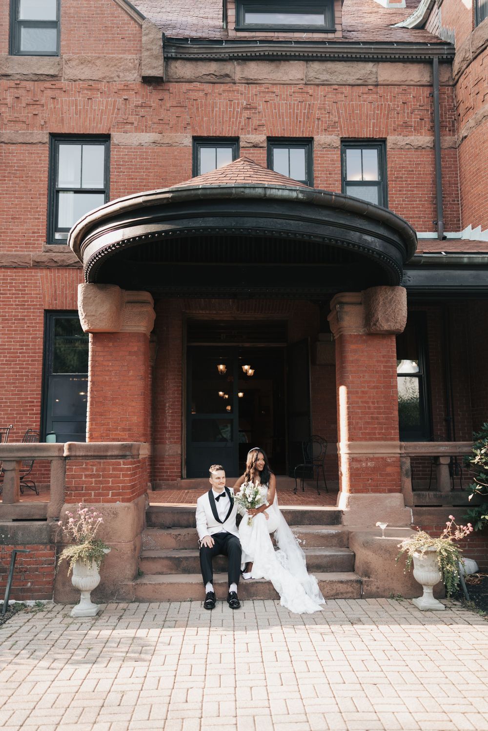 Bride and groom sitting on steps of elegant red brick building with rounded portico entrance on their wedding day.
