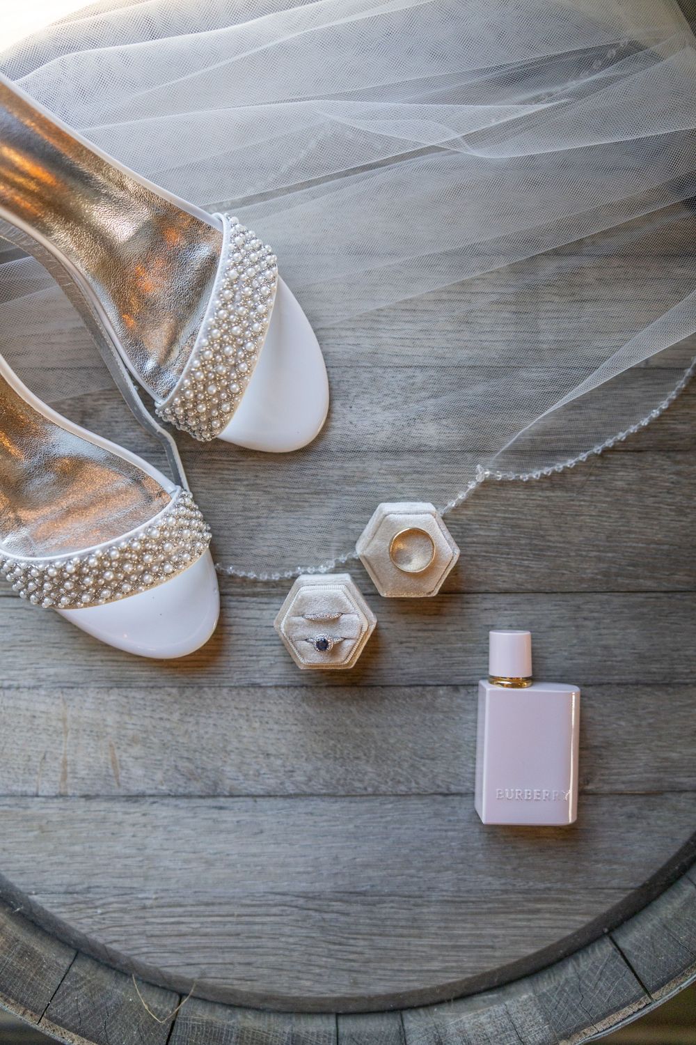 Wedding accessories including pearl-studded shoes and a hexagonal perfume bottle arranged on a rustic wooden surface.