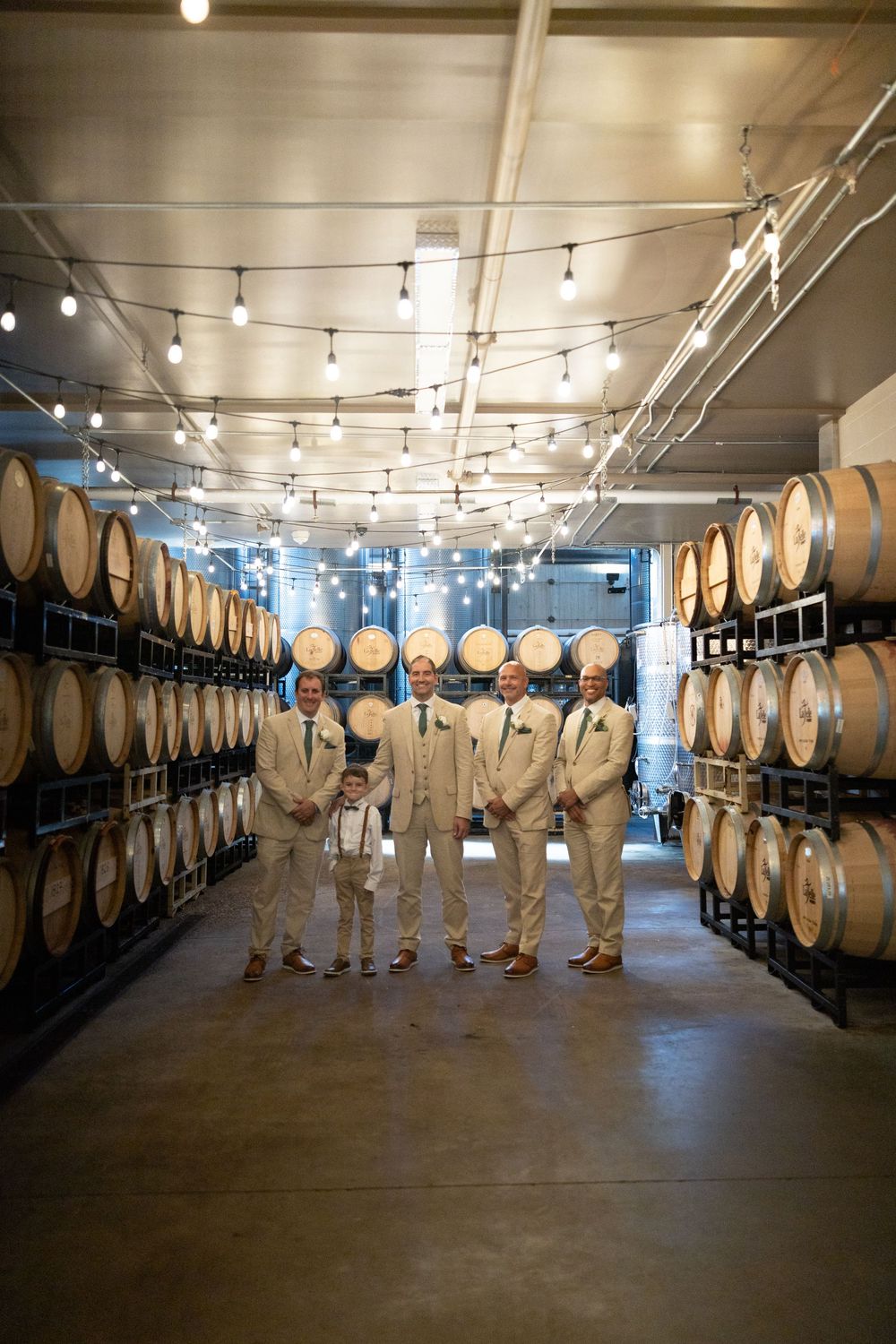 A group in light colored attire stands in a wine cellar with rows of wooden barrels along the walls.