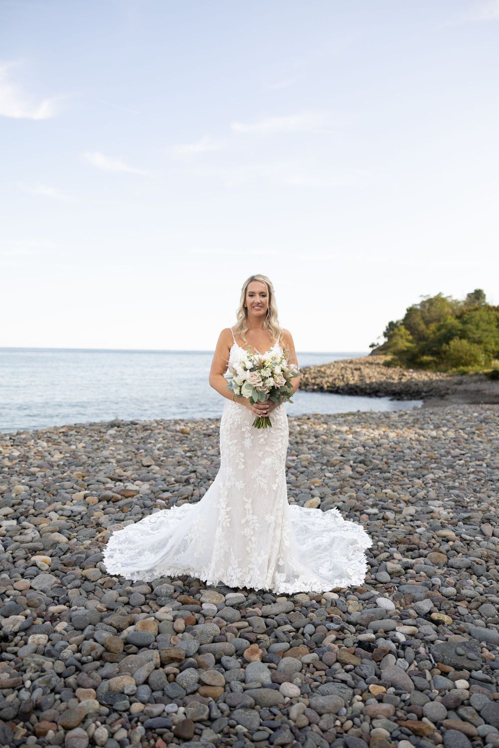 A bride in a lace wedding dress sits gracefully on a rocky beach shore with ocean waves and greenery in the background.