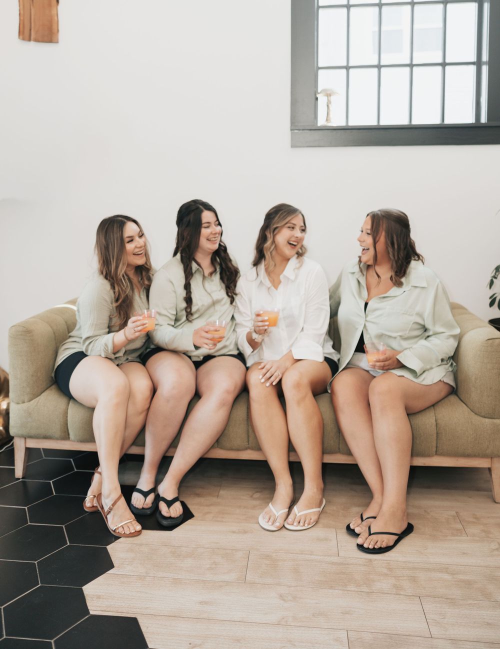 A group of friends wearing matching light colored outfits laugh together while sitting on a beige couch.